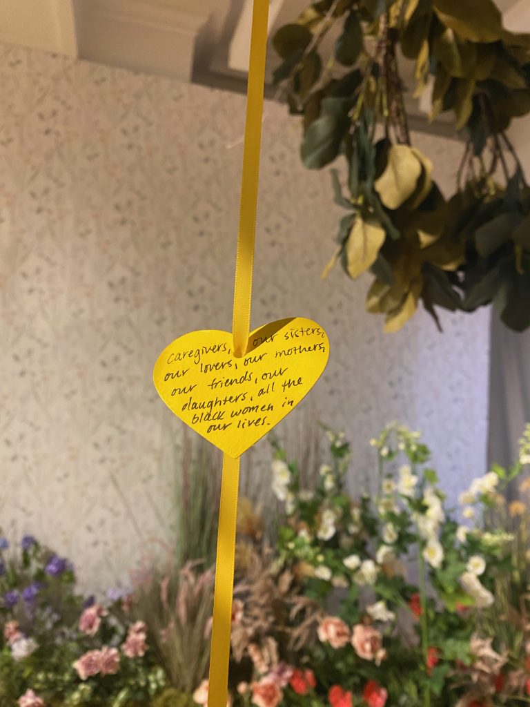 Image: "Wishes for Black Girls" hung on Rekia' Boyd's Ancestral Tree. A yellow ribbon strung through a yellow paper heart. Written on the heart in black cursive is the text "caregivers, our sisters, our lovers, our mothers, our friends, our daughters, all the black women in our lives." In the background are a variety of flowers and the branches of a fig tree. Photo by Jasmine Barnes. 