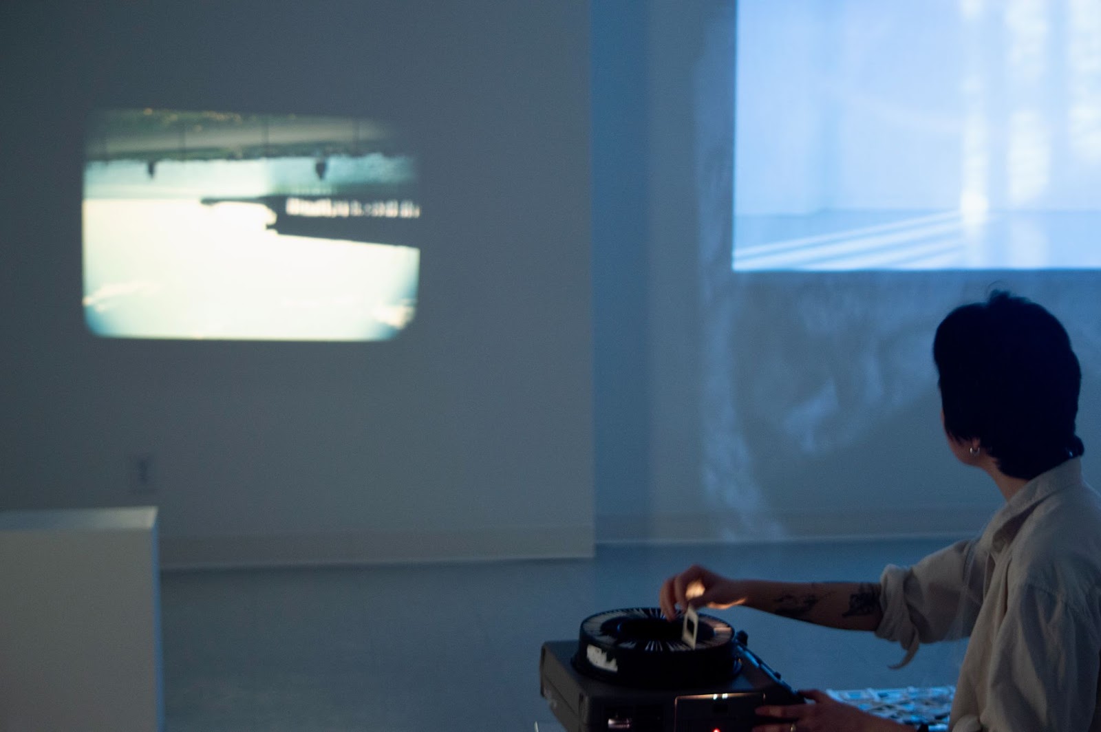 Image: A still of the closing performance for Ruby Que’s Consider a Disappearance. An image of an upside-down shoreline is projected onto a white wall. A light blue, still image of a video work is visible on the right side of the image. A figure crouches in the bottom right corner, inserting photo slides into a projector. Photo by Ethan Vergara