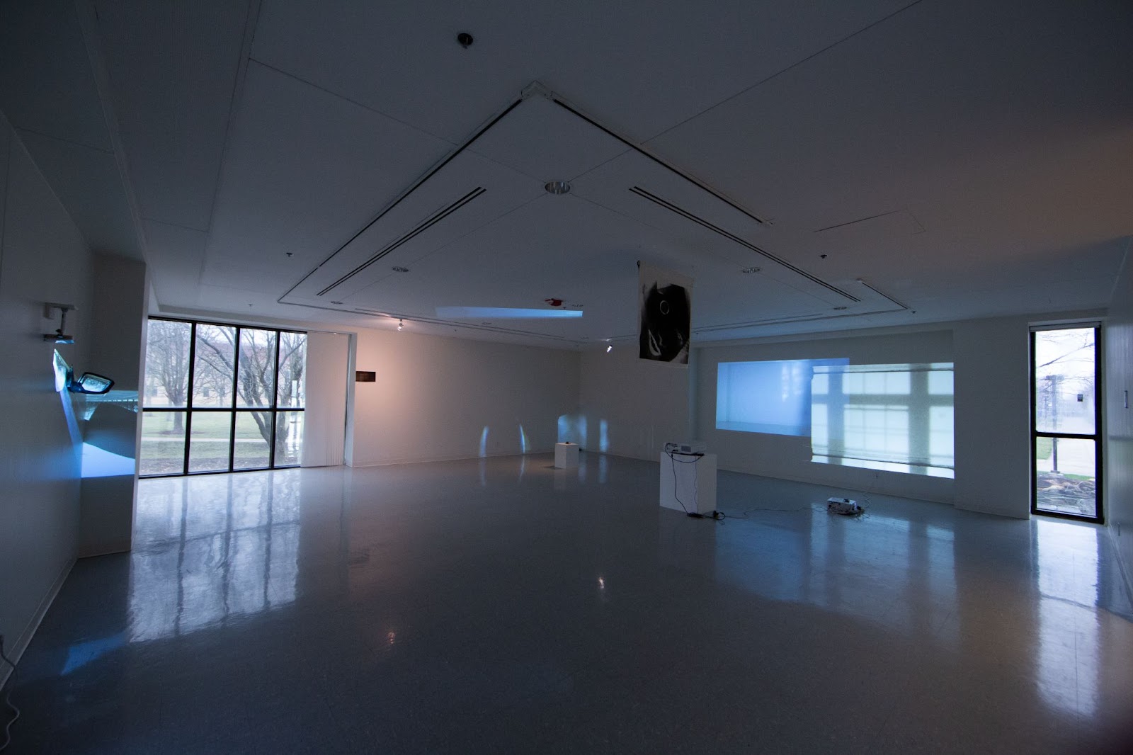 Image: Installation view, Ruby Que, Consider a Disappearance. Several projectors cast bluish, spectral light onto the walls of a dimly-lit gallery. A large paned window in the left corner of the room reflects on the floor. Photo by Ruby Que