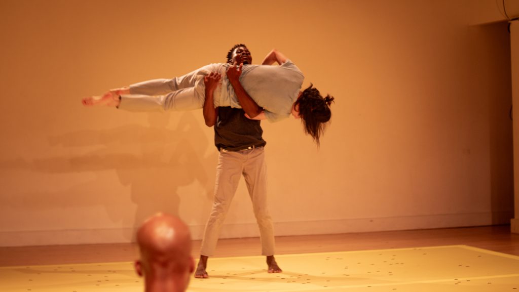 Image: A dancer holds another dancer horizontally in both his arms. Their shadows are cast softly and repeatedly on the wall behind them. The back of an audience member's head is in the foreground, next to the standing dancer's foot. Photo by Jovan Landry.