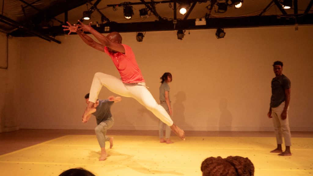 Image: A dancer wearing a salmon-colored shirt lunges in the air towards the left side of the image. Directly behind him is one figure near crouching and another standing, facing the back wall. To the right of the dancers in motion is one more dancer standing neutral, looking towards the camera. Photo by Jovan Landry.