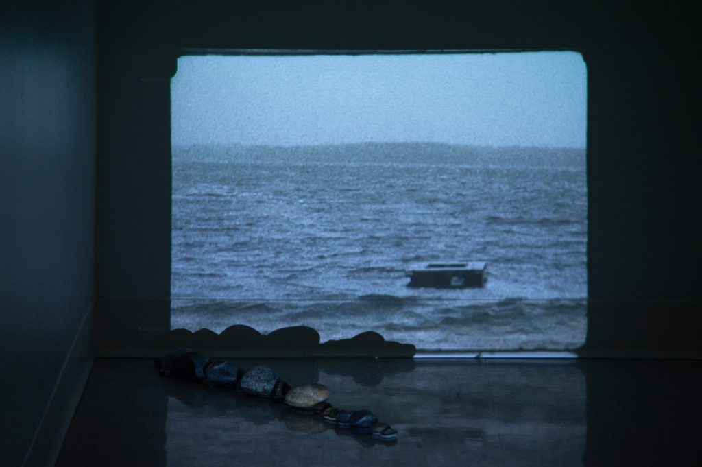 An image of a wooden platform floating adrift at sea is projected onto a wall. A line of stones is arranged in a diagonal along the floor, starting in the left corner and extending to the left side of the image. 