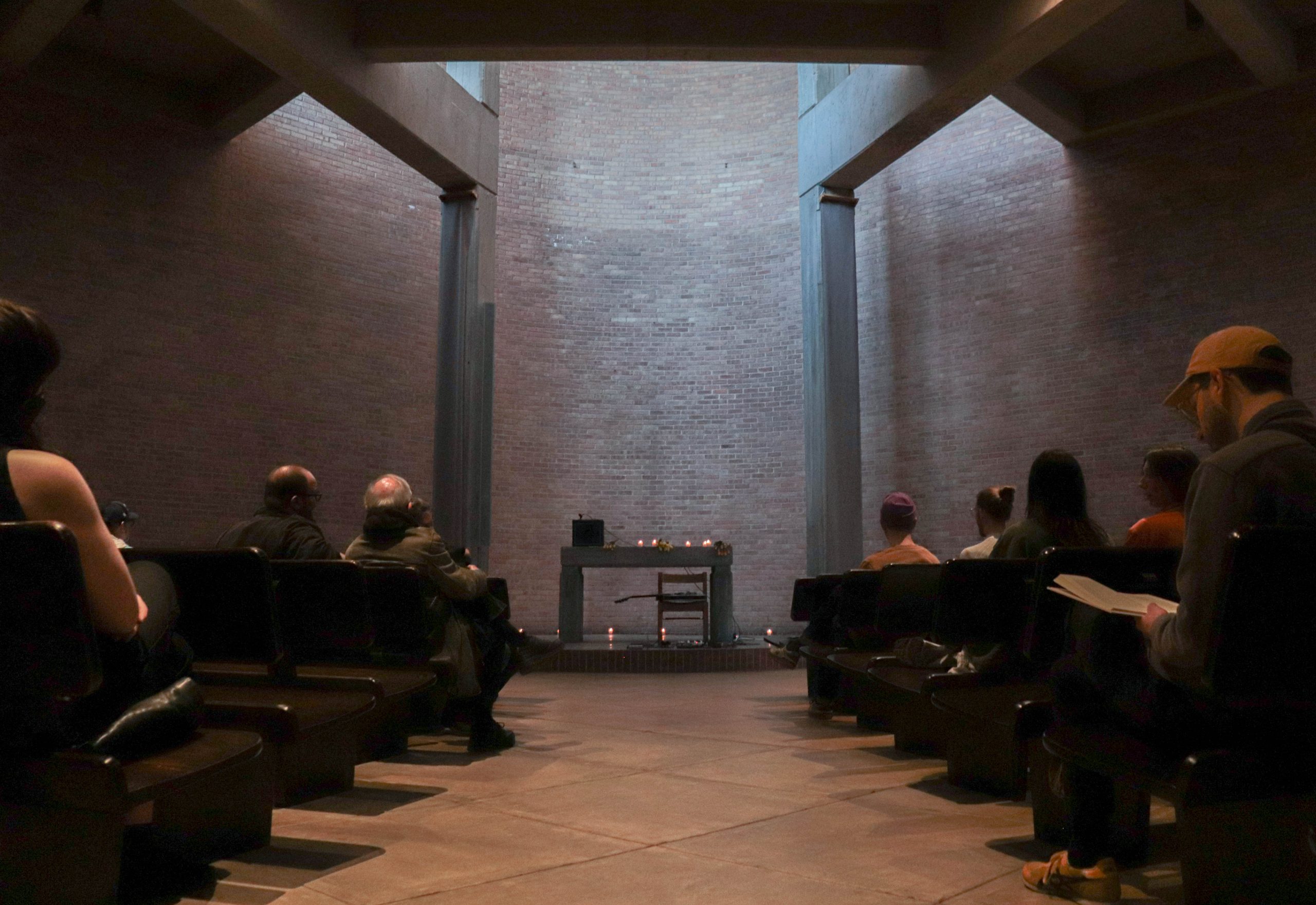 Image: People sit in the pews at Augustana Lutheran Church to participate in the Deep Listening Workshop. Light streams in from above, illuminating a small stage where a table and chair, candles, a guitar, and an amplifier are arranged as if waiting performance. Photo courtesy of Livy Snyder.