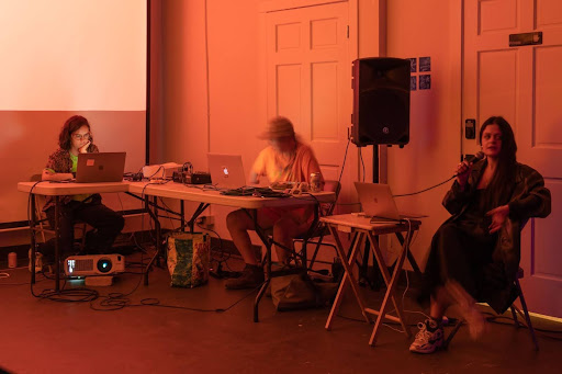 Image: Three seated people facing their laptops during Dark and Hot in front of the projector screen at Comfort Station. Photo by Jonas Mueller-Alheim, 2023.
