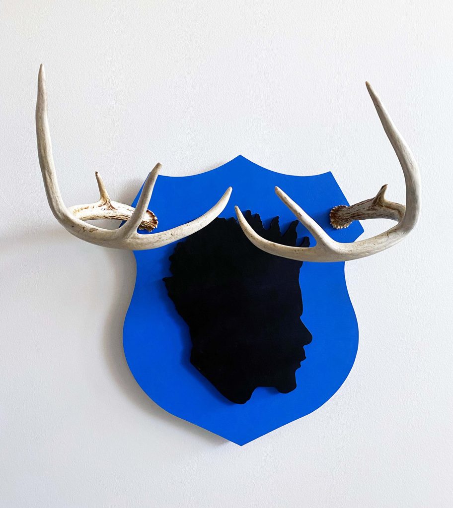 Image: Kevin Demery, Buck. 19x16x10. Fiberboard, acrylic, and whitetail deer horns. Silhouette of head in black centered on top of a blue badge. Antlers are placed on either upper corner of the blue badge. Image courtesy of Gallery Bogart.