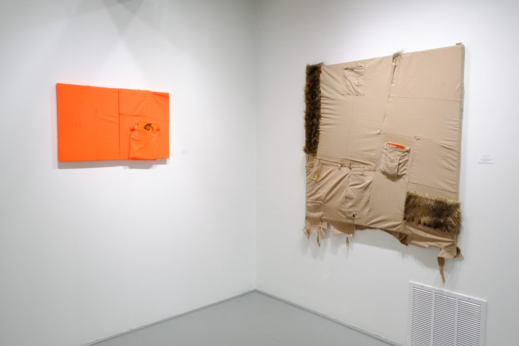 Image: Gallery view of two works hung near opposing corners on the white walls of the gallery. Left: Andrew Mcilvaine, I Am, Am I, Aim, 30 x 32". Laser-etched, bright orange hunting overalls and vest stretched over canvas with "Mexican-Americans South of Texas" book in pocket. Right: Andrew Mcilvaine Worn in & Stretched Out, 48 x 48". Tan dickies, animal fur, collage, gold grill, and antlers mounted to canvas. Image courtesy of Gallery Bogart.