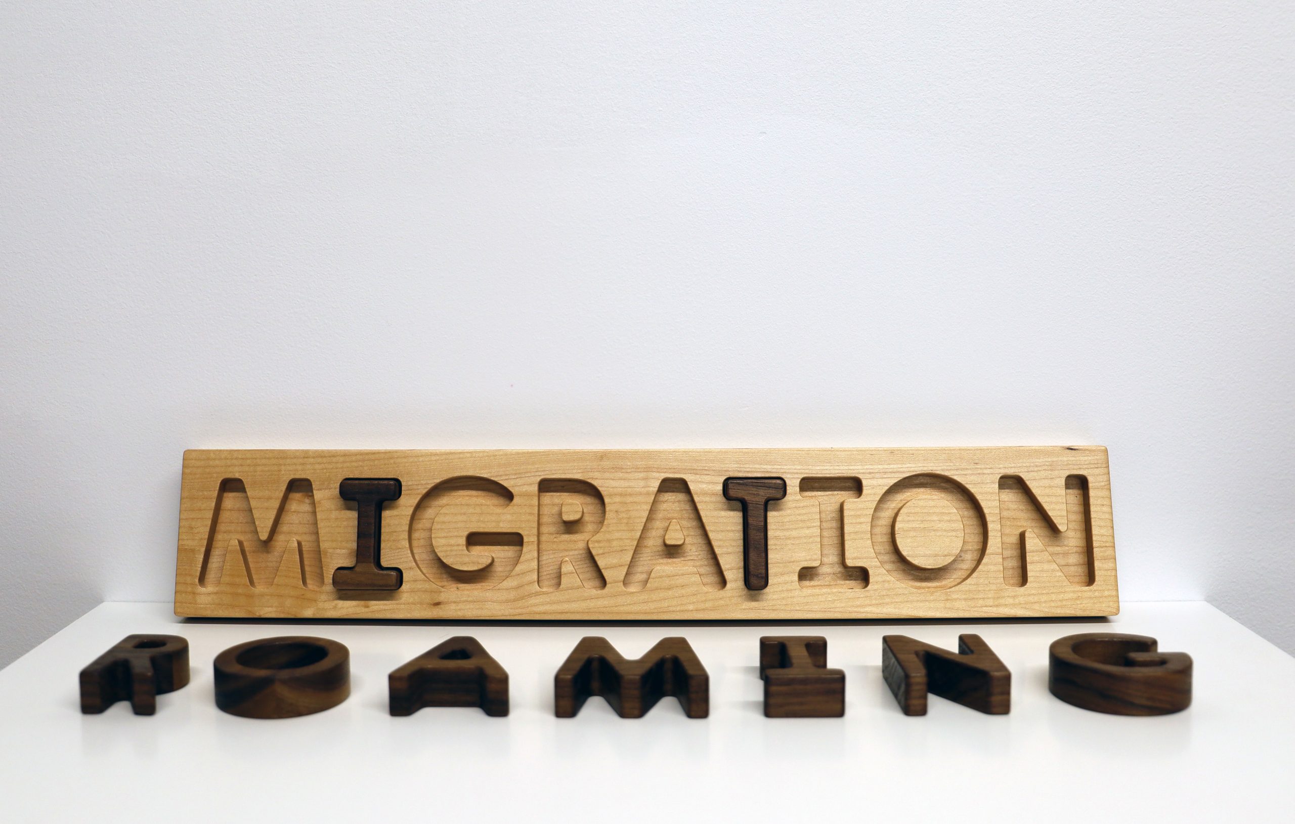 Image: View of Kevin Demery's Migration, oak and walnut wooden puzzle, 3.5 x 19". The word "MIGRATION" is lasercut on a rectangular block of light-colored wood and shown leaning on a plinth against the gallery wall. The word "ROAMING" is shown laid flat on the plinth using dark-colored wood cut-outs. The "I" and "T" in "MIGRATION" are filled in using dark-colored wood cut outs. Image courtesy of Gallery Bogart.