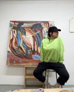 Image: Diana Motta seated on a stool in her studio, looking to the right of the photo. Behind her is her painting Inanna's Descent. She wears black pants, a black ball cap, and a fluorescent green sweater.