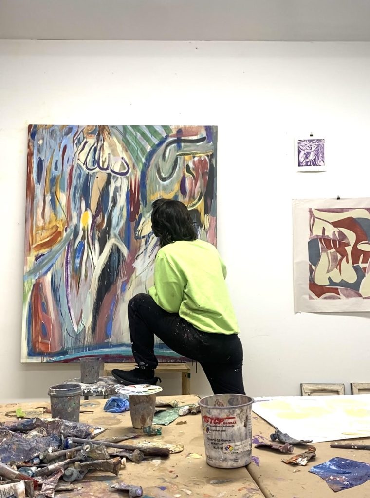 Image: Diana Motta in her studio. She wears a fluorescent green sweater and black leggings, facing away from the camera, towards a painting that she works on Her left foot is placed on a stool. To her right is a smaller piece of canvas on the wall. Behind her, closer to the camera is a table, covered with discarded paint supplies, cups, and other detritus. Image courtesy of the artist.