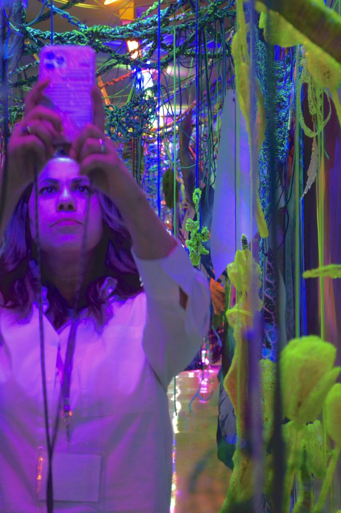 Image: A phantasmagoria of fibers and yarn in black light blues and purples and highlighter yellows, hanging from the ceiling and knit into cables that stretch across the room. In the forefront of the image is a person, lit purple, wearing a white button-up shirt. They're focused on the phone they're holding up above and before their head. Image by Luz Magdaleno Flores.
