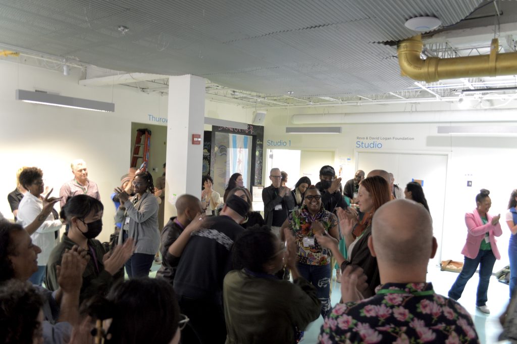 Image: In a large white-hued room, about 20 people mill about with their hadns in various forms of gesticulation: some appear to be clapping others, hold their fingers in a state of waggle. Others are smiling and patting each other on the back. In the back of the room are two doors with the words "Studio 1" and "Studio" above them. Image by Luz Magdaleno Flores.