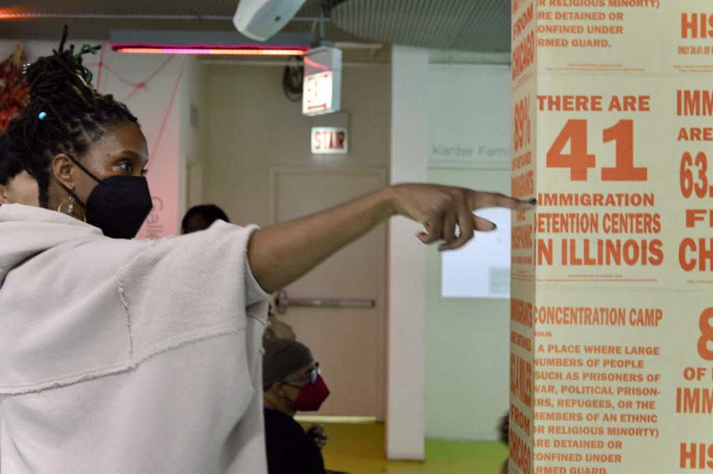 Image: A Black person wearing a grey shawl and a black N95 face mask with their hair up, points their hand up and their finger to the right of the image. On a beige pillar where their finger ends is orange text describing facts about US policies towards immigrants. The text their finger is in-line with reads "THERE ARE 41 IMMIGRATION DETENTION CENTERS IN ILLINOIS." Behind the person are other people, seated and standing. There is a door and an exit sign in the background. Image by Luz Magdaleno Flores.