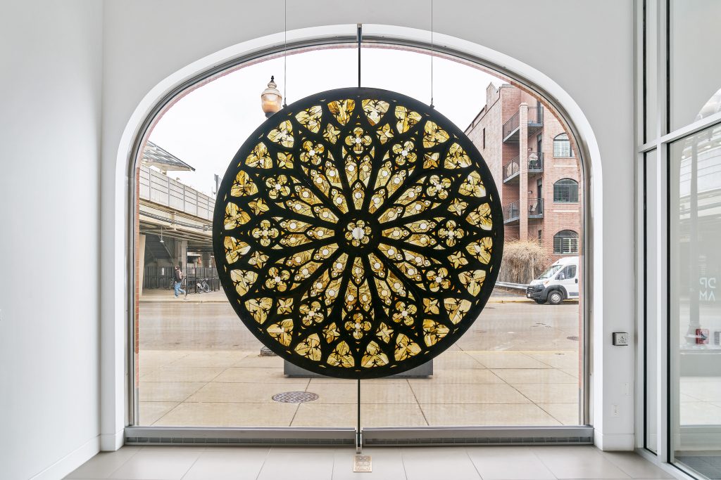 Image: Selva Aparicio, Remains, 2023-24, discarded lettuce from 2013, steel, oil board, and UV plexi. In Memory Of, installation view, DePaul Art Museum. Courtesy of the artist. An installation piece that looks similar to a a rose window, an architectural element found in Gothic cathedrals, displayed upon a glass wall. Photo by Bob.