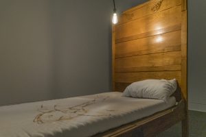 Image: Selva Aparicio, My Bed, 2023-24, twin size bedframe and mattress, bed sheet, Betadine, and suture. Courtesy of the artist. In Memory Of, Installation view, DePaul Art Museum. A bed with no blankets and a wooden headboard sits in a gallery. One single naked bulb lights the space above the bed. The sheets of the bed are stained. Photo by Bob.