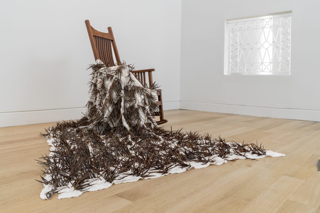 Image: In Memory Of, installation view, DePaul Art Museum. Selva Aparicio, Saudade, 2023, Simax glass window bars and temporary wall. Selva Aparicio, Solace, 2023-24, crochet cotton blanket and honey locust thorns. Courtesy of the artist. A crocheted blanket is draped over a wooden rocking chair. The blanket is filled with sharp, protruding honey locust thorns. Behind the chair on the right is a cut out of the wall that acts as a window into the next space. Image by Bob.