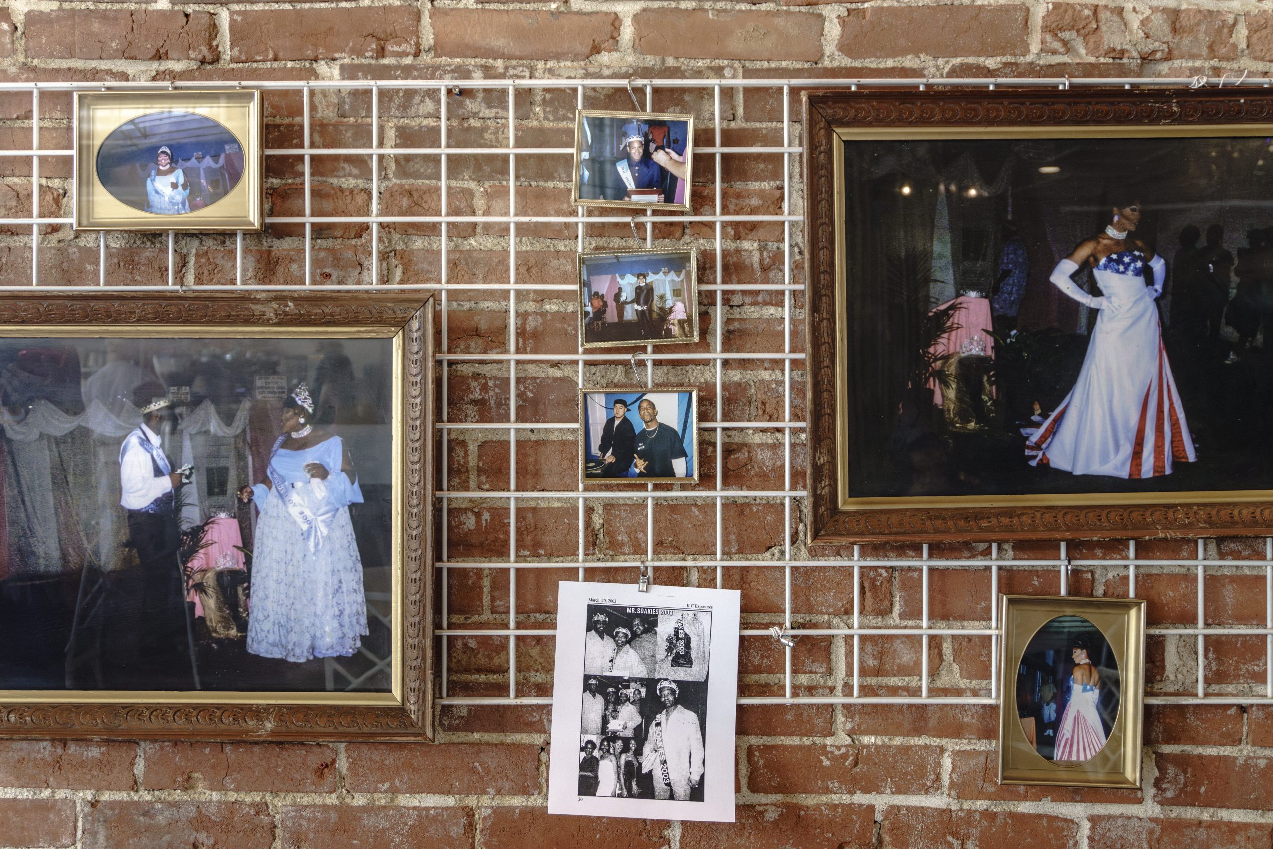 Image: Installation View, {B/qKC}: The Volume_2 Archive Launch Party. Archival photographs of queer pageant culture set on a brick wall. There are two large framed photos in the bottom left and the upper right of pageants. Photo by Gabriella Salinas.