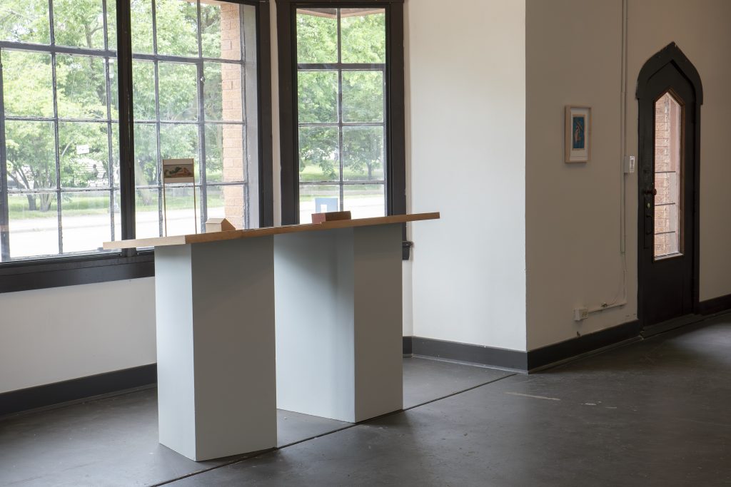 Image: Installation view of certain places at Comfort Station. To the left is Sheldon Till–Campbell's three-dimensional piece You are here, which is situated on two pillars next to a bay window, outside of which are a street and trees. Hanging on a wall to the right is a framed work on paper, next to a small wooden door with an arch and a window set in it. Photo by JI YAng. Instagram: @mr_he_he_he. Website: www.jiyang.co.