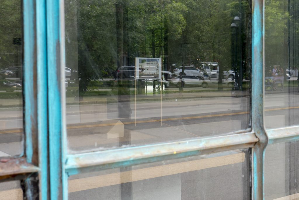 Image: Installation view of the exhibition certain places at Comfort Station, taken from outside the building looking in through a window. Through the window is the part of Sheldon Till–Campbell's piece You are here that looks like a billboard. Reflections of trees and cars can be seen on the window. Photo by JI YAng. Instagram: @mr_he_he_he. Website: www.jiyang.co.