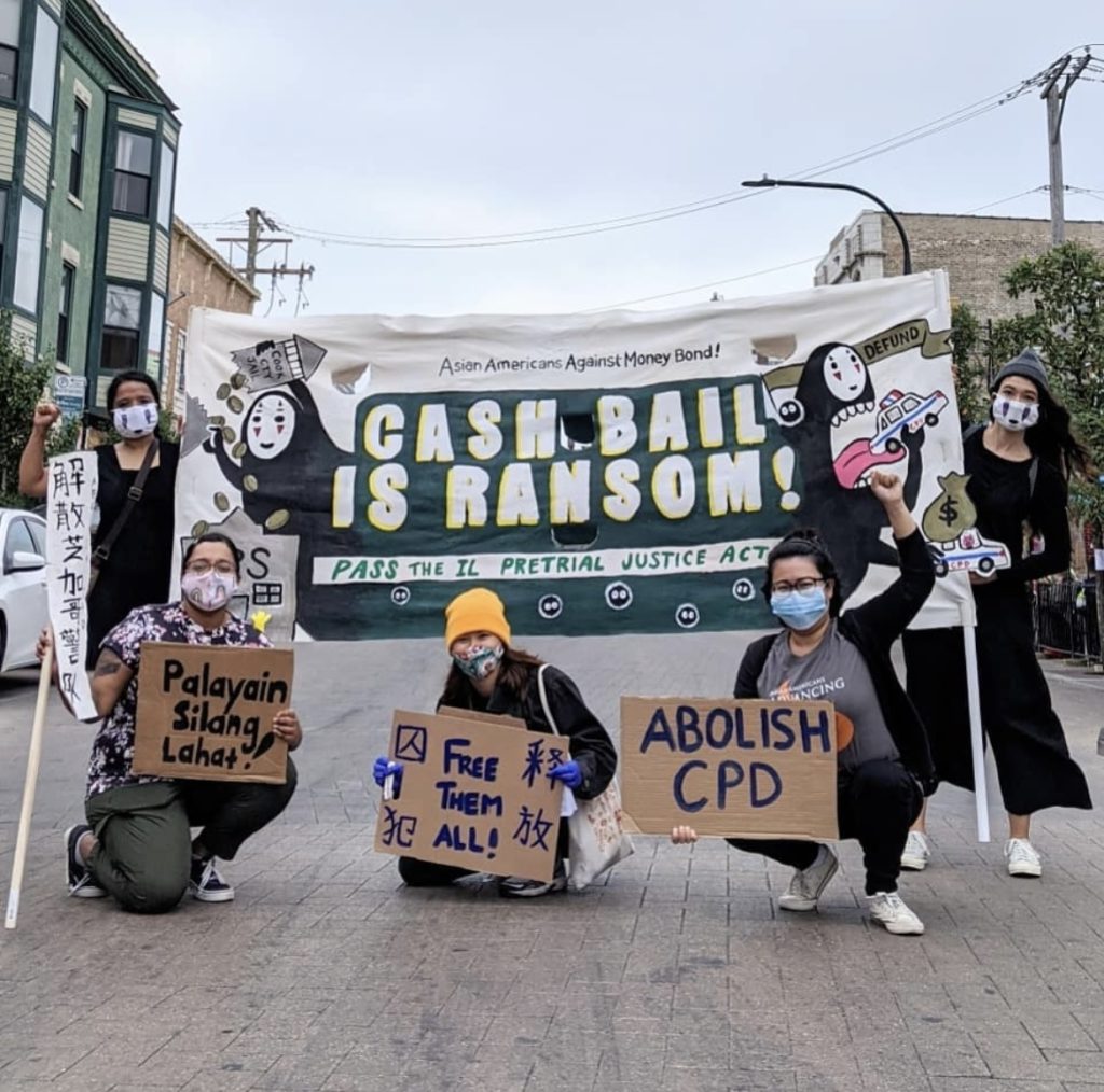 Image: Five members of A Just Chi and Nikkei Uprising pose for a photo holding signs and banners. The main banner, held by two individuals, reads "CASH BAIL IS RANSOM!" with smaller text "Pass the IL Pretrial Justice Act" and "Asian Americans Against Money Bond!" Other signs in the image include: "Palayain Silang Lahat!" (held by a person on the left), "Free Them All!" (held by a person in the center), and "ABOLISH CPD" (held by a person to the right of the center). The participants are wearing masks and standing on a street, with a mix of buildings and streetlights in the background. "Cash Bail is Ransom" banner designed by Cori Lin. Photo by aiko hamamoto.