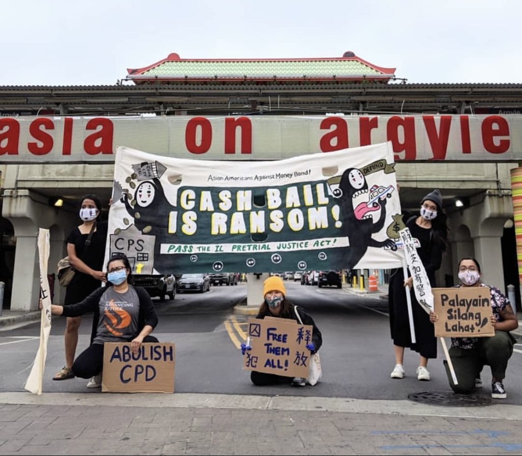 Image: Five members of A Just Chi and Nikkei Uprising, holding signs and banners. in front of a building with the signage "asia on argyle" visible above and behind them. The signs they hold include: "Palayain Silang Lahat!" (held by a person on the right), "Free Them All!" (held by a person in the center), "ABOLISH CPD" (held by a person to the left of the center). The participants are wearing masks and standing on a street, with a backdrop that includes a building with Asian architectural elements. Cash Bail is Ransom banner designed by Cori Lin. Photo by aiko hamamoto.