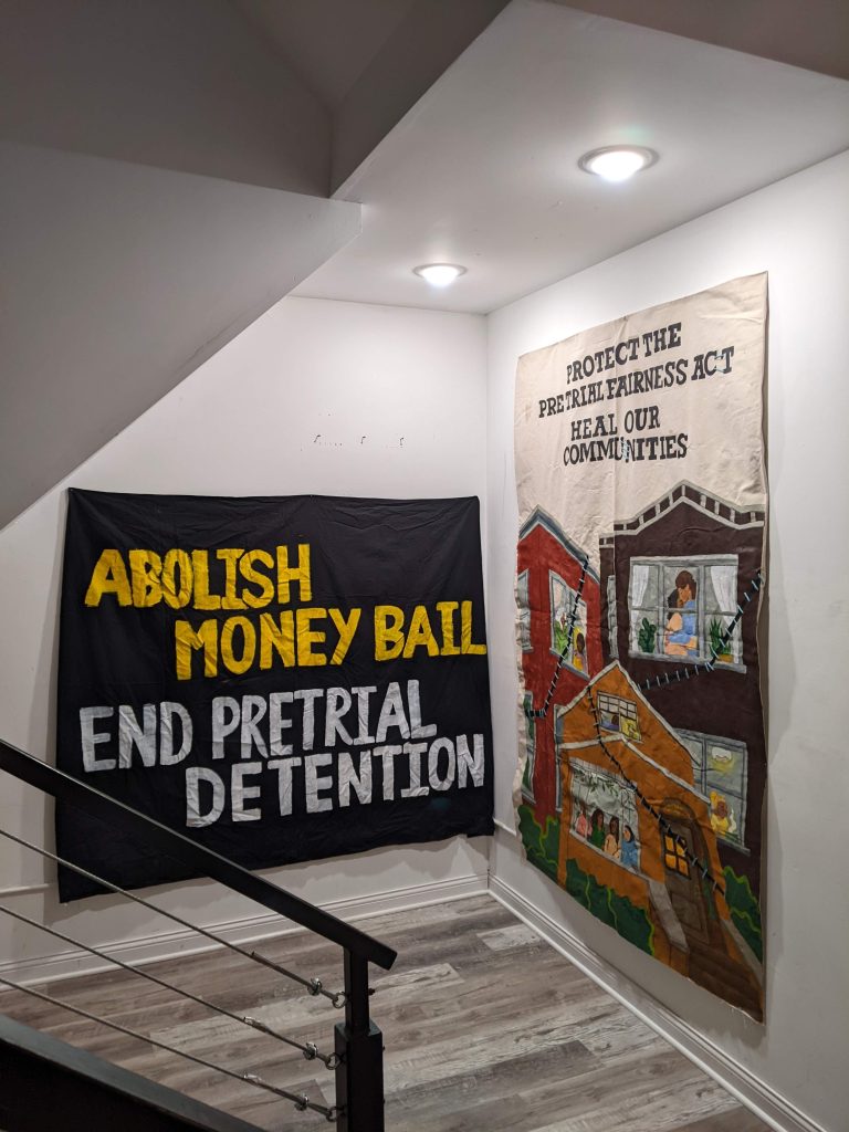 Image: Two wall-length banners in a stairwell as part of the Art in Action Exhibition. On the left is a black cloth banner with the words, "ABOLISH MONEY BAIL" and "END PRETRIAL DETENTION" on it. Designed by INPJ network. To the right on different wall, is a banner with three houses on it with people hugging and standing next to each in the windows. The houses each of a stich running across them. At the top, there is text which reads "PROTECT THE PRETRIAL FAIRNESS ACT. HEAL OUR COMMUNITIES." Designed by Kara Rodriguez. Photo courtesy of the author.