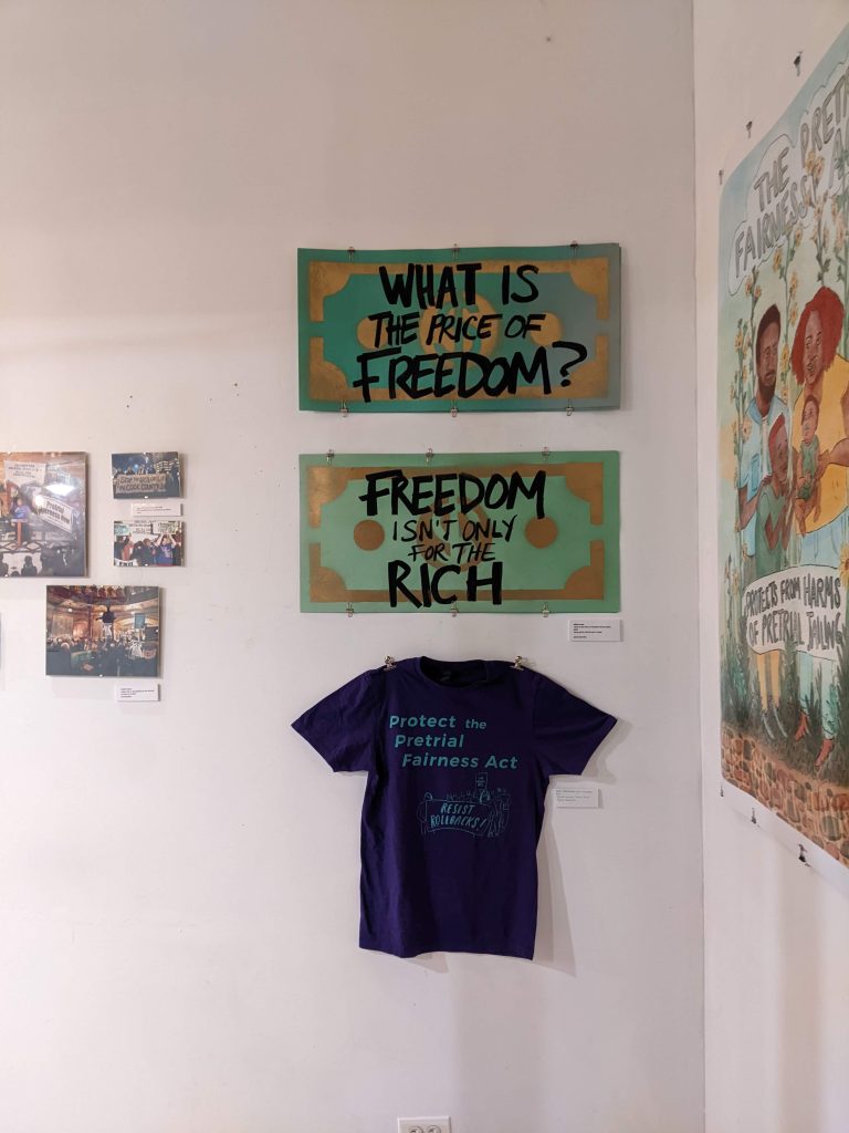 Image: Photo of eight art pieces as part of the Art In Action Exhibition, including: large dollar signs with green backgrounds and gold accents;,one reads "WHAT IS THE PRICE OF FREEDOM?" in bold black letters and the other dollar sign with a similar design reads "FREEDOM ISN'T ONLY FOR THE RICH" in black letters. Made by Olivia Love. Below these signs, a purple T-shirt hangs, displaying the message "Protect the Pretrial Fairness Act" in turquoise letters. Created by Cori Lin based on an illustration by Danbee Kim. To the left are four photos of key protests advocating for the Pretrial Fairness Act. On the right side of the display, part of a large poster is visible, depicting illustrations and text that reads "THE PRETRAIL FAIRNESS PROJECT" and "PROTECT FROM HARMS OF PRETRIAL JAILING." Photo courtesy of the author.