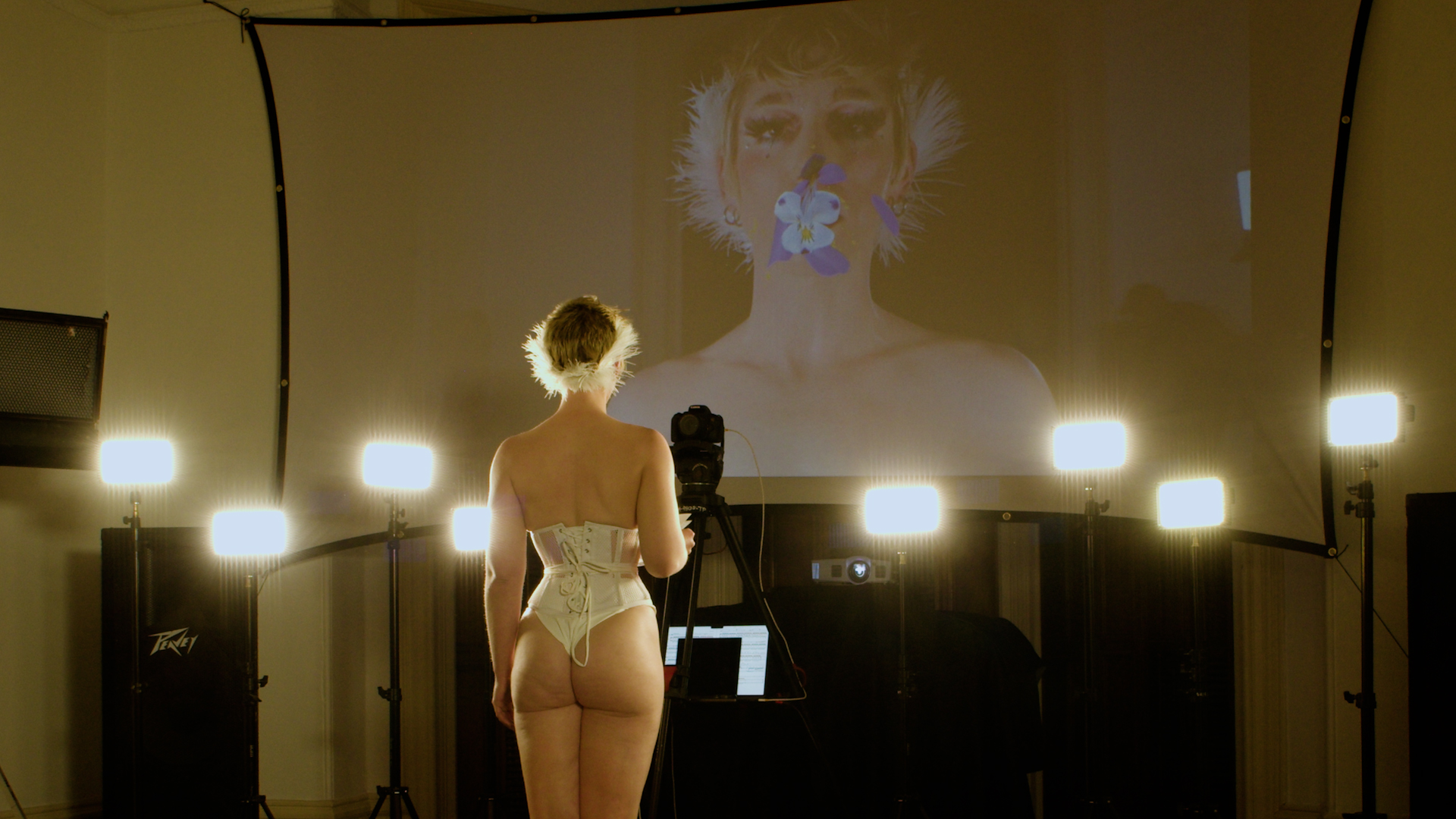 Image: Bun Stout performs “The Swan” at PS1 Close House. A performer wearing a white corset and underwear poses in front of a projector screen with their back turned toward the camera. Their arms are lifted above their head and are flexing, displaying the rippled muscles of their back. A line of unlit lights is arranged in front of the projector screen. A luminous sphere is displayed on the projector screen. Photo courtesy of the artist.
