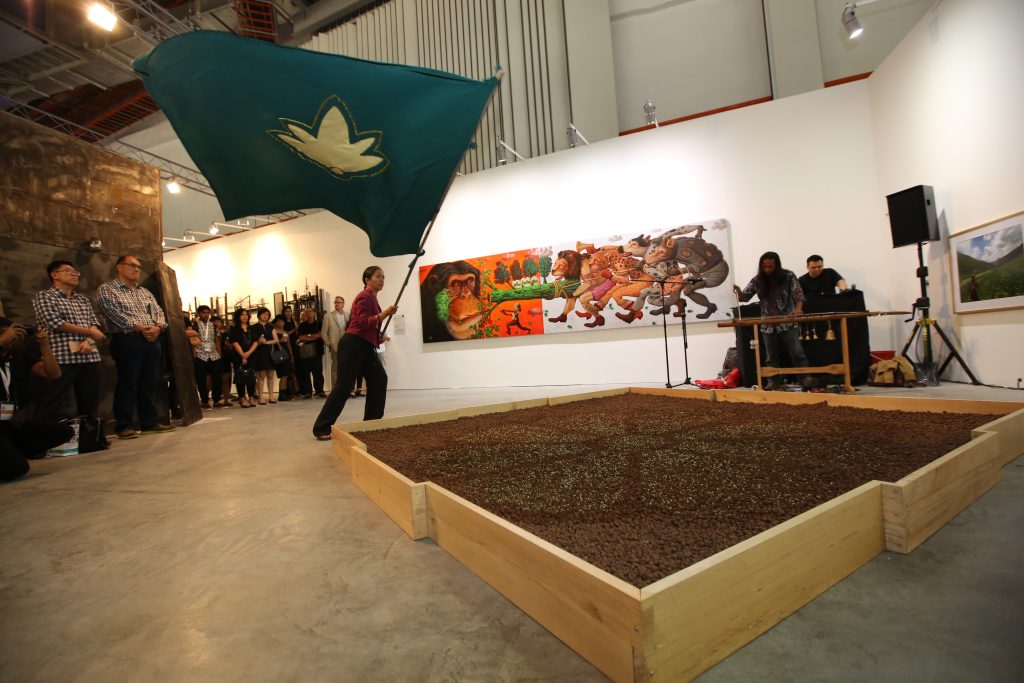 mage: Arahmaiani, Memory of Nature, 2013, Singapore. During a performance in a gallery space, Arahmaiani raises a large teal and gold flag around a bed of seeds which are patterned to look like a flower. There are behind her and two muscians playing bells and other instruments to her left. A painting depicting the struggle of domestication versus nature and a photograph of a lush green landscape are line the walls of the gallery across from the onlookers. Courtesy the artist.