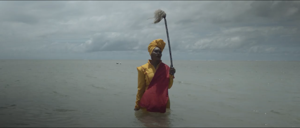 Image: Lhola Amira, IRMANDADE: The Shape of Water in Pindorama, 2018-2020, HD video, single
channel sound, film still. Amira is facd towards the camera. She stands leg-deep in the ocean wearing a yellow and burgundy outfit and yellow turban. She is carrying a gray staff or wand which may have spiritual and cultural significance. There is a breeze and light waves in the water. Image courtesy of SMAC Gallery, copyright Lhola Amira.