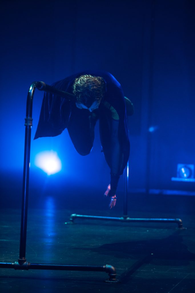 Image: Performance still of "Memories of a Supernatural AIDS Crisis" in Detroit. A person lying on a horizontal ballet barre facedown, their legs and arm draped towards the ground. Behind them, a blue light glares at the camera.. Photo by Erika Ruch. Courtesy of Marc Arthur.