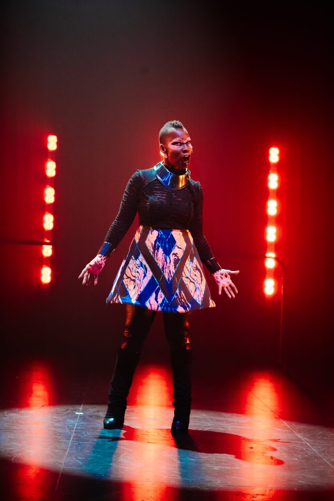 Image: Performance still of "Memories of a Supernatural AIDS Crisis" in Detroit. A person stands with their arms outstretched slightly from their sides, their palms face up. Their mouth is wide open and their brow is furrowed; their expression is accentuated by white makeup across their eyes. Behind them,  two rows of flaring red LED lights. Photo by Erika Ruch. Courtesy of Marc Arthur.