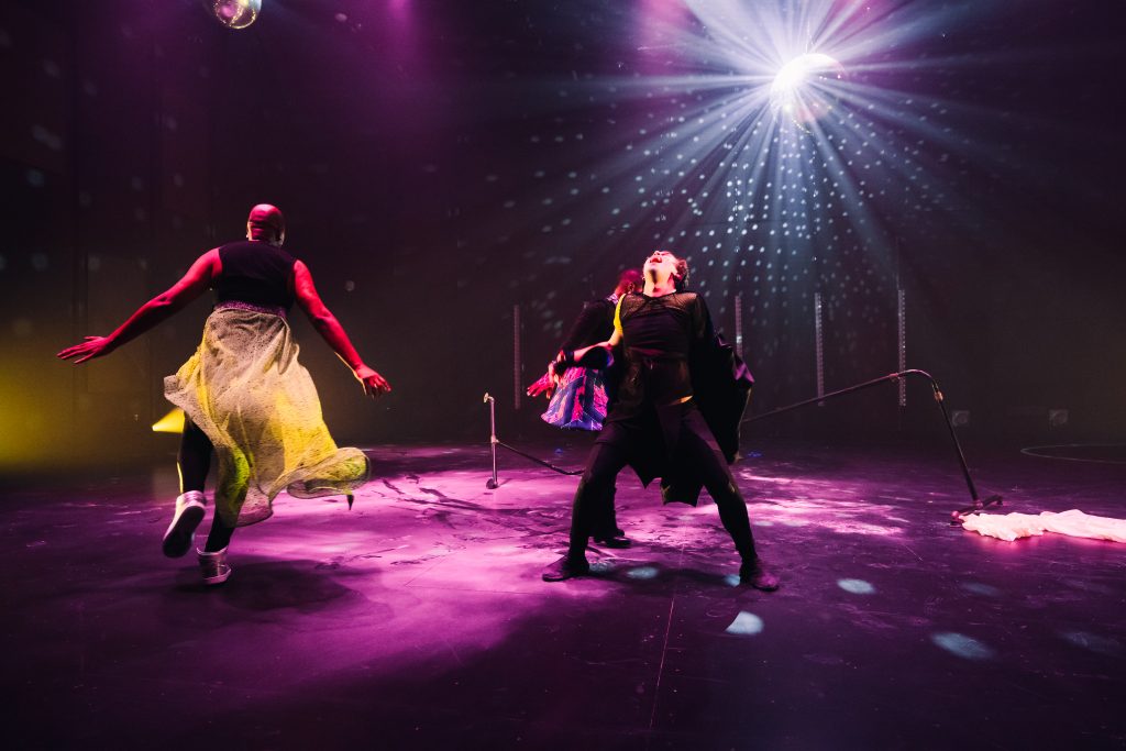 Image: Performance still of "Memories of a Supernatural AIDS Crisis" in Detroit. The ballet barre is broken, and a disco ball gleams from the background casting a pink and spotted silver light onto the wall stage floor, as three performers dance: the one to the left, with their arms out to their side, skips away from us; the one to the right throws their head back with their legs spread; the third performer is obscured behind the second. Photo by Erika Ruch. Courtesy of Marc Arthur.