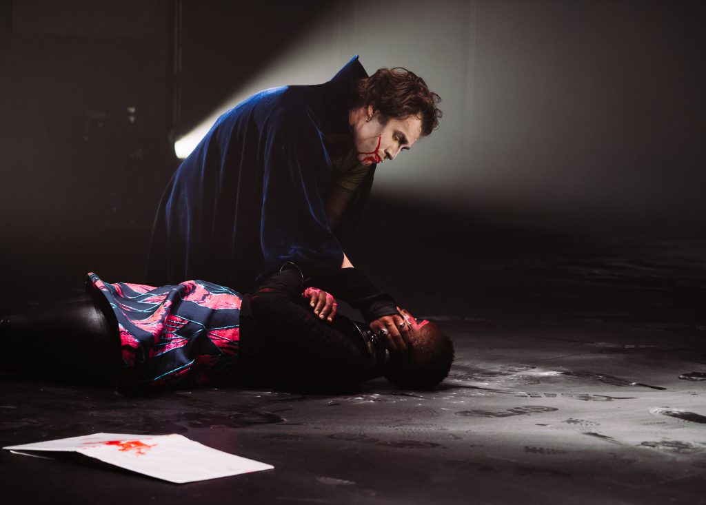 Image: Performance still of "Memories of a Supernatural AIDS Crisis" in Detroit. An actor with a gash on their cheek and a long blue cape kneels down over another performer lying on the ground. The first performer has placed their hand across the other's mouth. The floor is dusty with footprints across it. In front the performers on the floor is a white piece of paper with a red smatter on it. Photo by Erika Ruch. Courtesy of Marc Arthur.