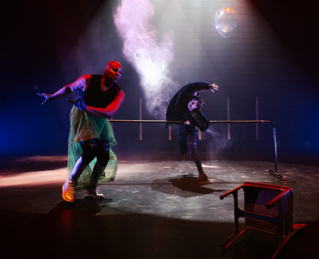 Image: Performance still of "Memories of a Supernatural AIDS Crisis" in Detroit. A moment of movement where in the background a spotlit person leans against the ballet barre and raises an arm over their head as smoke rises behind them. The person in the foreground leans forward. To the right of them is a chair on the ground. There is a disco ball hanging from the ceiling.  Photo by Erika Ruch. Courtesy of Marc Arthur.