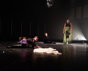 Image: Performance still of "Memories of a Supernatural AIDS Crisis" in Detroit. On a mostly bare black stage, three performers. One on the left wearing pink-eye make-up is stretched across the floor, on their knees, reaching for a white cloth in the center of the stage. One in the back sits cross-legged staring up at the one on the right, who stands, looking down towards the first. Behind the first is a fallen chair. Behind the standing figure is a white spotlight. There is a disco ball hanging from the ceiling. Photo by Erika Ruch. Courtesy of Marc Arthur.