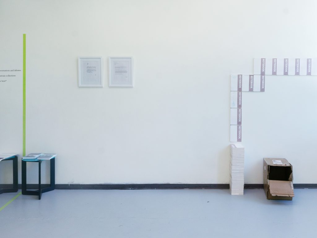 Image: Wang Langgou, Instructions, 2017, (left) and Forced Intersection of Mediums, 2017-2022 (right). An installation view of On Acknowledgements / Lines of Handling at State VIII Project. A line of green tape on the wall and floor on the left side of the image separates the room and two tables with publications. Two framed sets of instructions are hung in the center of the wall. An empty box with a flap cut out sits on the right side of the image. A stack of notebooks sits to the left of the box and a line of notebooks are displayed on the wall above the notebook and box. Photo by Tianjiao Wang.