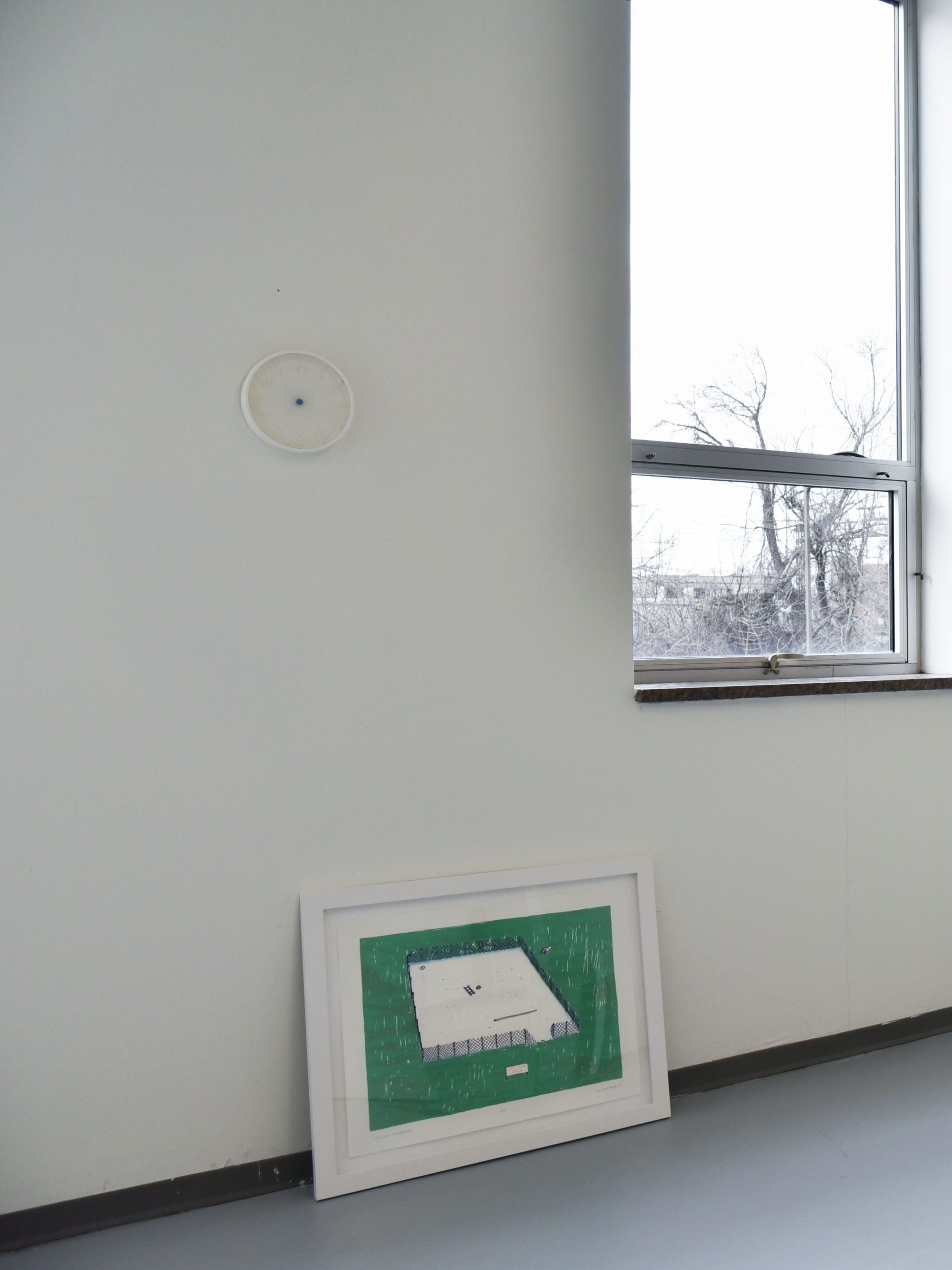 A framed print of a playground in the center of a green field sits on the floor of a studio and is propped against the wall. A small IKEA clock is hung on the wall above the print. There is a window to the right of the clock. 