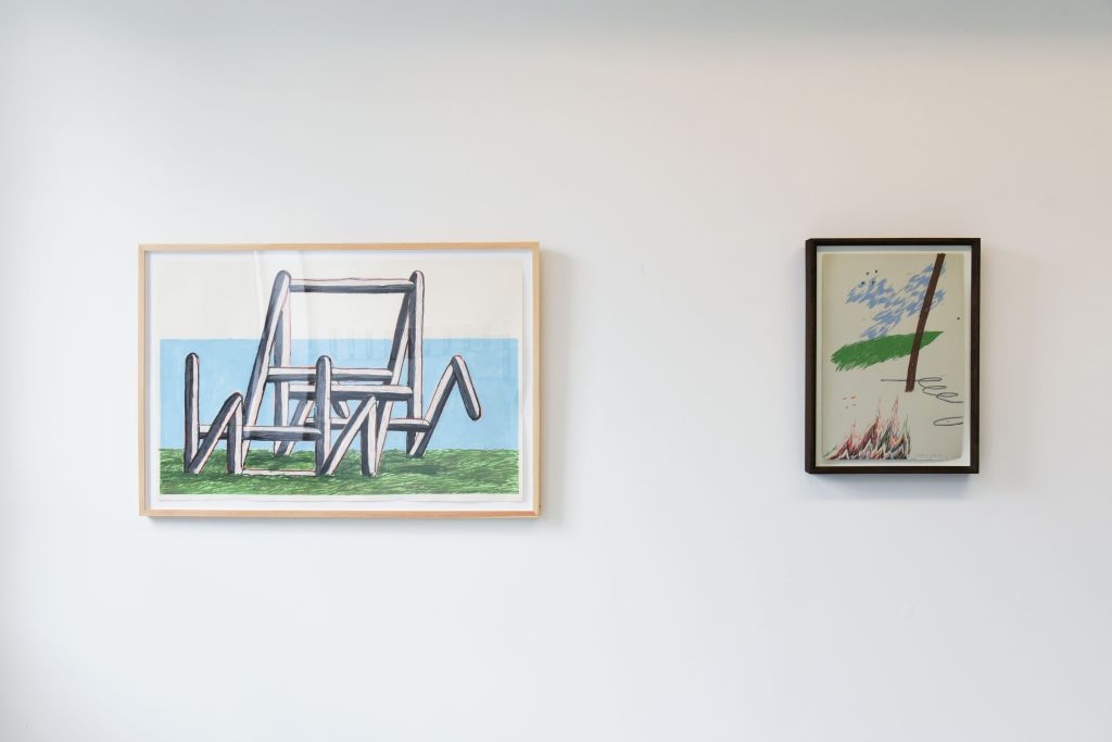 Image: McKinzie Trotta, Ten-foot Ladder, 2021 (left) and Put it Out, 2022 (right.) Two framed prints are hung on a gallery wall. The left pictures a geometric structure made from sticks resting on a green horizon against a blue and white background. The right is a smaller scene of a cross-shaped blue sky floating above a green horizon with a hose snaking from the right side of the image and ending behind a brown pole. A multicolored fire rises from the bottom of the image. Photo by Tianjiao Wang.