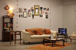 Image: Installation view, People Who Came Before Us. In the center is a large, low coffee table on a red patterned rug, a beige couch with caramel-colored throw pillows and a patterned throw. To the left of the couch, there is a television on a side table. On the wall behind the couch is a gallery of vintage photos featuring Ifti Nasim and groups of other South Asian LGBTQ+ figures. There are colorful decorative garlands with shapes of elephants cut out. On the left side of the wall is a shelf with books, CDs, a framed photo, and a lit table lamp. Courtesy of South Asia Institute.