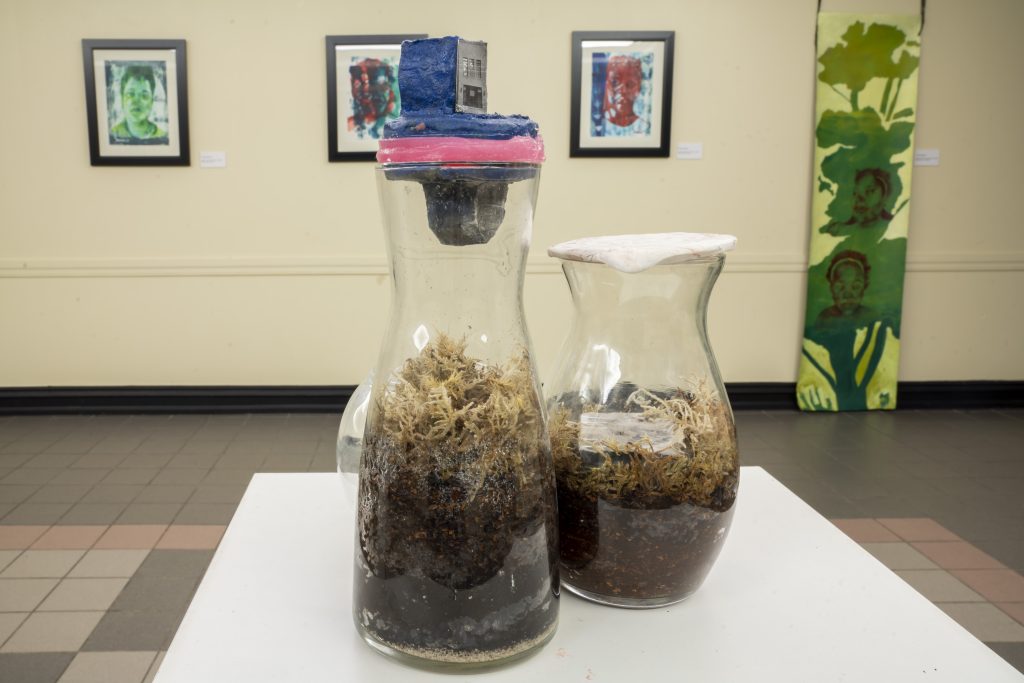 Image: Ghetto Terrariums Series: Meditation on What Persists, by Tavia David, installed as part of Finding Ceremony—Tiffany Johnson's project for her 2022 Anchor Curatorial Residency at Austin Town Hall. Two glass vessels sit on a pedestal with organic matter inside. In the background, several works of art hang on a wall: three depicting figures and one vertical piece in greens and yellows. Image courtesy of Tiffany Johnson. Photographer: Vanessa Lara/ Colectivo Multipolar.