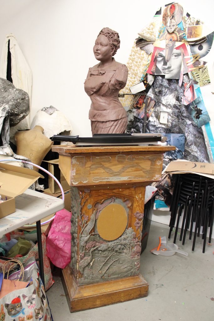 Image: A view of Sonja Henderson's studio, with various collage elements, a bust of a woman, and art supplies behind a wooden podium. Photo by Prince Jai.