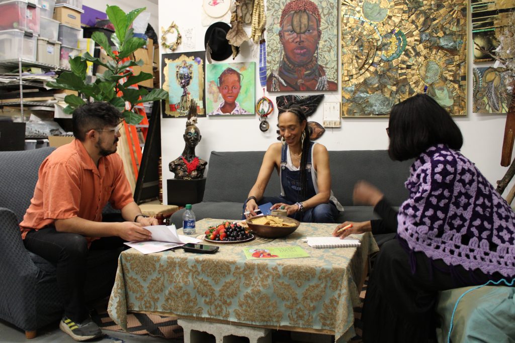 Image: (from left to right) Carlos Flores, Sonja Henderson, and Tiffany Johnson sit in Henderson's studio around a table. Behind the three are various works of art by Henderson hung on a wall. Photo by Prince Jai.