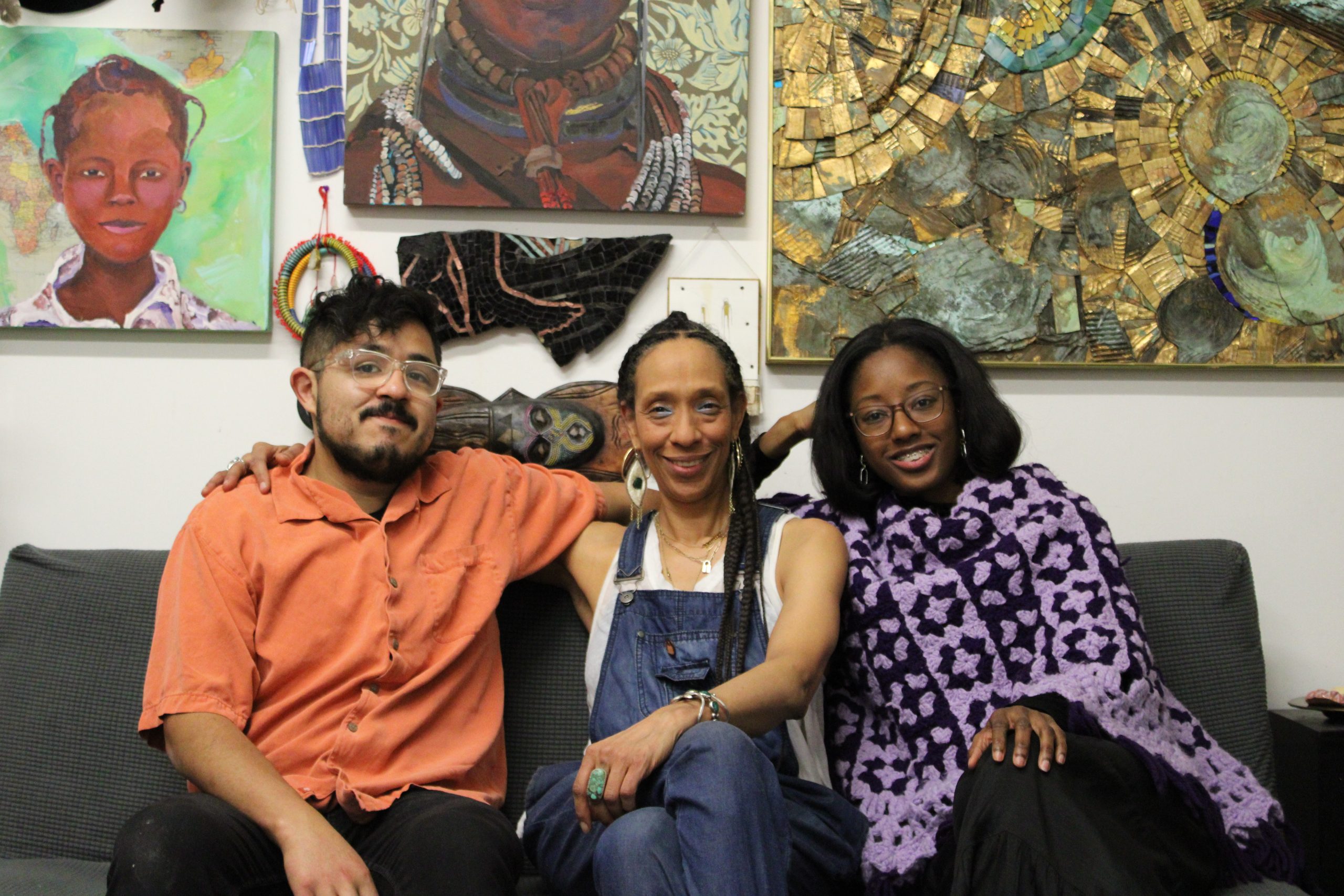 Image: (from left to right) Carlos Flores, Sonja Henderson, and Tiffany Johnson sit on a couch in Henderson's studio smiling at the camera. Behind the three are various works of art by Henderson hung on a wall. Photo by Jai Prince.