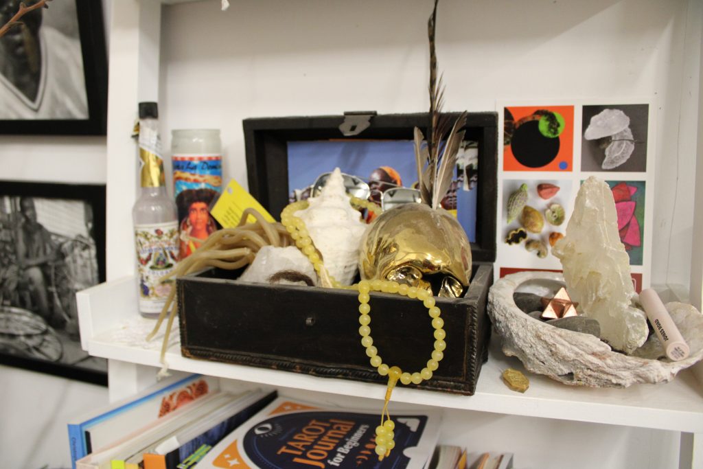 Image: A view into Sonja Henderson's studio. A box of different trinkets, such as a shell, beads, feathers, and a golden skull, sits on a shelf next to other objects like a crystal and a candle. Surrounding the shelf are photos and books. Photo by Jai Prince.