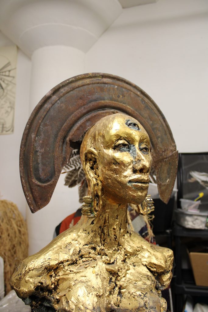 Image: A gold bust by Sonja Henderson in her studio with a metal, halo-like headpiece. The bust has a third eye and is facing to the viewer's right. Photo by Prince Jai.