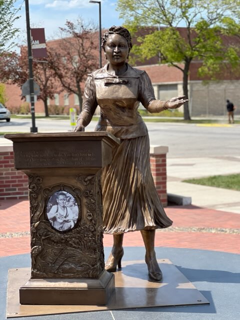 Image: The Mamie Till-Mobley and Emmett Till Memorial by Sonja Henderson. The memorial is comprised of a woman (Mamie Till-Mobley) standing facing the viewer with one arm extended out. The other arm touches a podium with ornate flowers and plants that surround an image of Emmett Till on the base. Image courtesy of the artist.