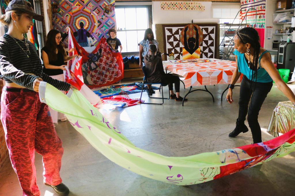 Image: A gathering of the Mother's Healing Circle organized by Sonja Henderson. A group of women hold various textile piece, many of them being colorful quilts. Image courtesy of Sonja Henderson.