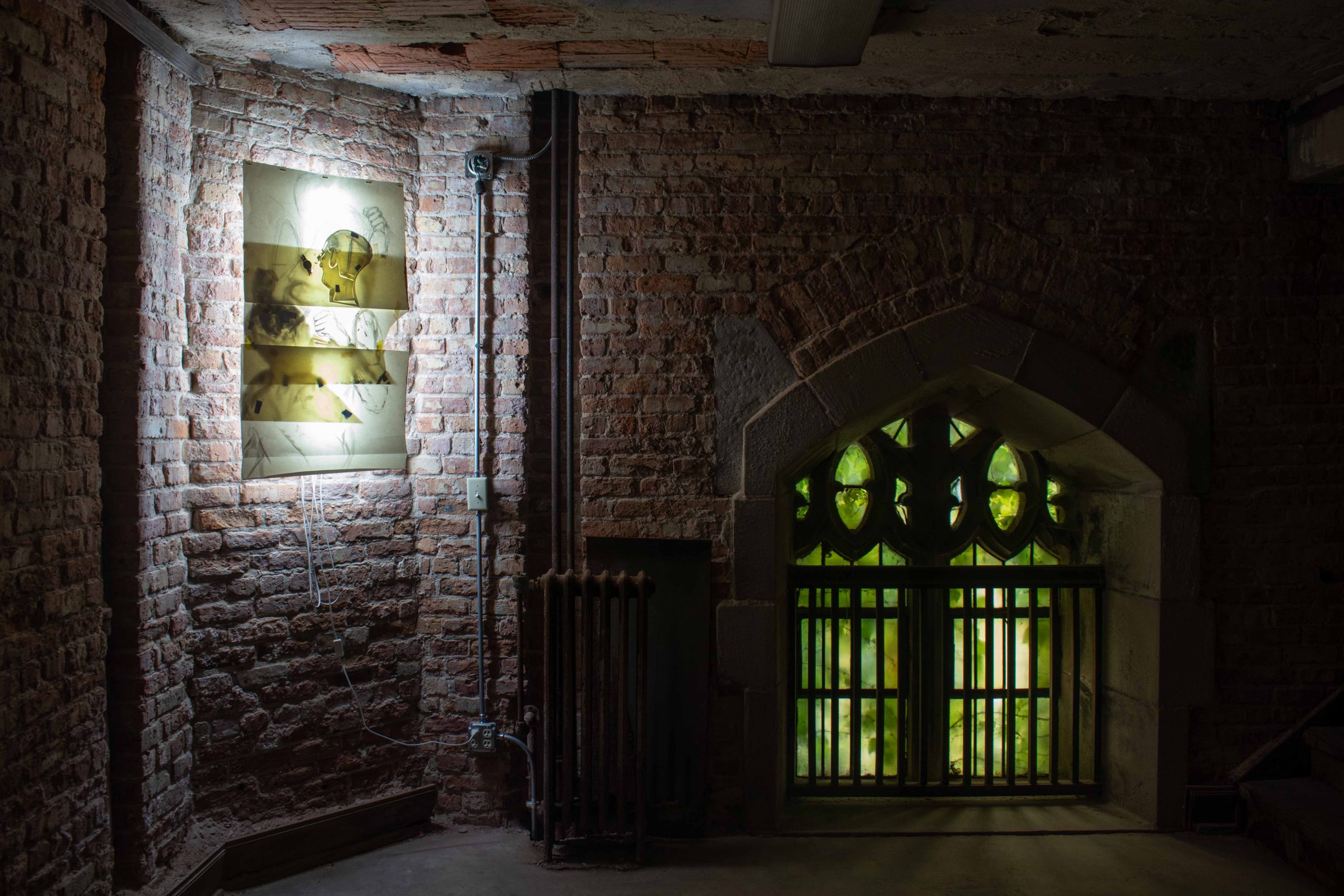 Image: Installation view of the World Friendship Room on the second floor of the First Presbyterian Church of Chicago. A drawing is lit from the back and hung in a dark room with a wall of exposed bricks and pipes. The drawing consists of three taped pieces of paper depicting three figures lining vertically engaging in some violent exchanges. A physical rainbow-colored wristwatch is worn by the figure on the top. An arch-top window sits on the ground right to the drawing and a staircase goes up towards the right. Photo by Tianjiao Wang.