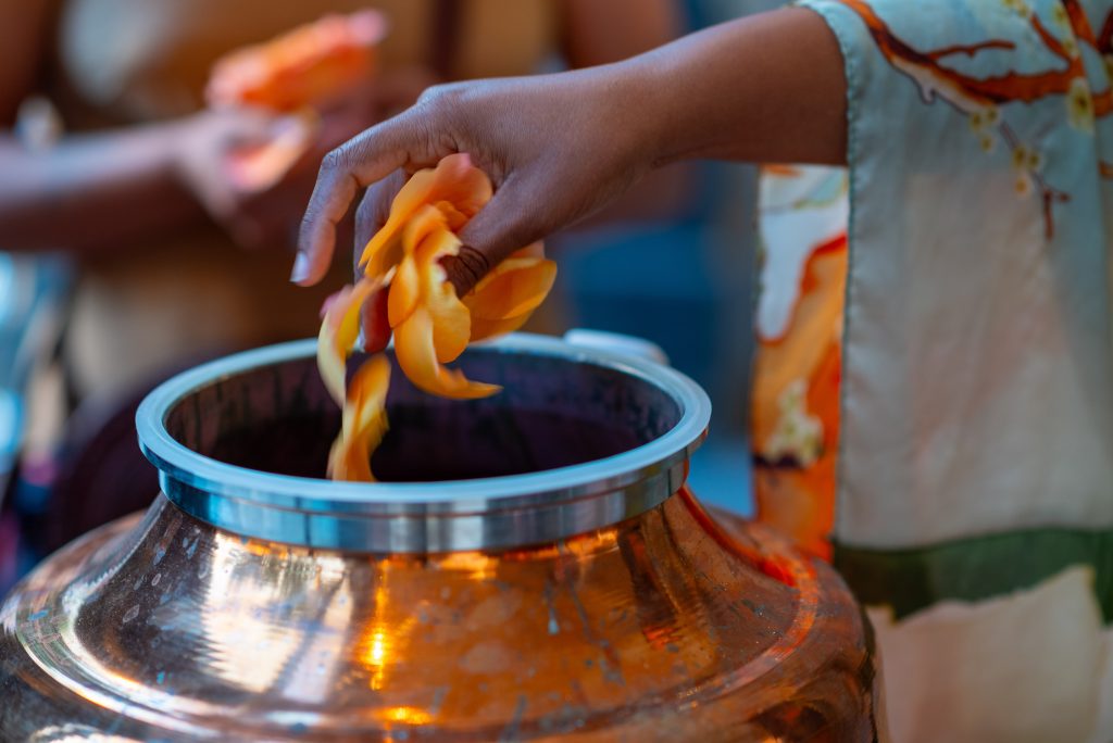 Image: A hand releases orange rose petals into a copper distiller. The arm releasing them has a robe draped over it. Photo by Tonal Simmons.