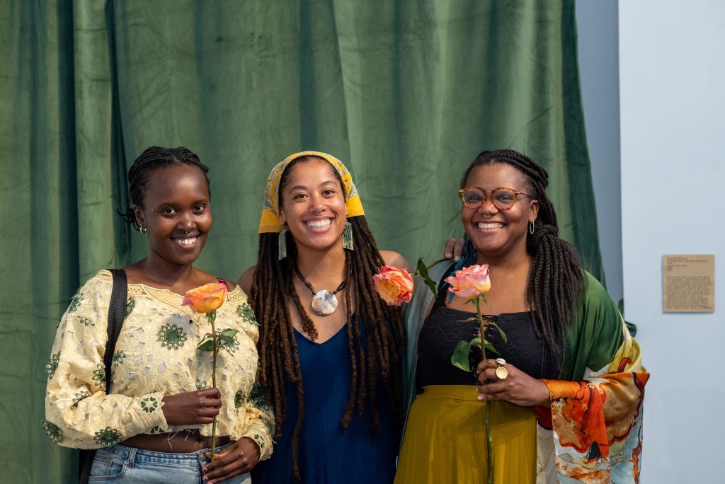 Image: From left to right, Evéa, Courtney Morrison, and Atoi Glennette, co-creators of the film come alive, smile and hold orange-pink roses on the opening night of piecemeal. Behind them is an olive green curtain. Photo by Tonal Simmons.
